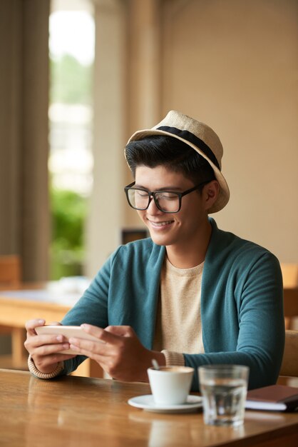 Lächelnder stilvoller asiatischer Mann, der im Café sitzt und Mitteilungen auf Smartphone überprüft