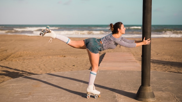 Lächelnder tragender Rollschuh der jungen Frau, der auf einem Bein am Strand balanciert