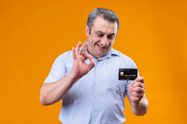Lächelnder und positiver Mann mittleren Alters im blau gestreiften Hemd, das Kreditkarte betrachtet, während ok Handzeichen auf einem orange Hintergrund zeigt