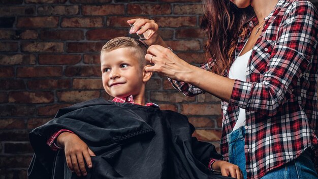 Lächelnder Vorschuljunge, der einen Haarschnitt bekommt. Kinderfriseur mit Schere und Kamm schneidet kleinen Jungen im Raum mit Loft-Interieur.