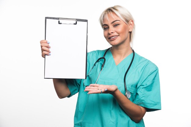 Lächelnder weiblicher Arzt, der eine Zwischenablage hält, die Hand auf weißer Oberfläche zeigt