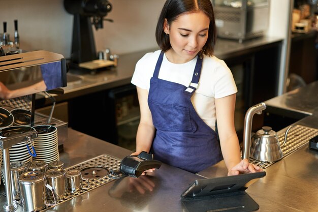 Lächelndes asiatisches Barista-Mädchen, das im Café an der Theke arbeitet und Bestellungen über den POS-Terminal-Empfang bearbeitet