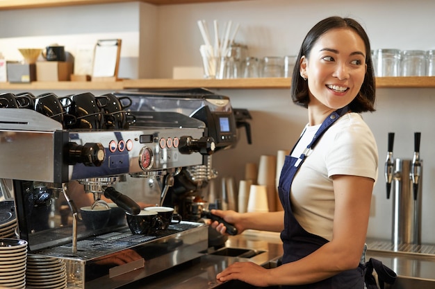 Lächelndes asiatisches Barista-Mädchen macht Cappuccino mit Kaffeemaschine steht hinter der Theke im Café