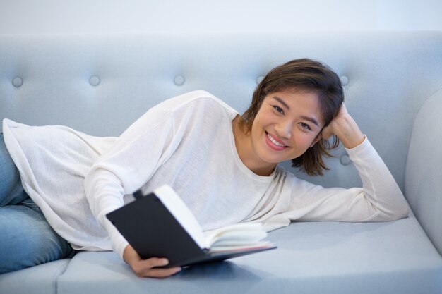 Lächelndes asiatisches Mädchen, das auf Trainer liegt und Tagebuch liest