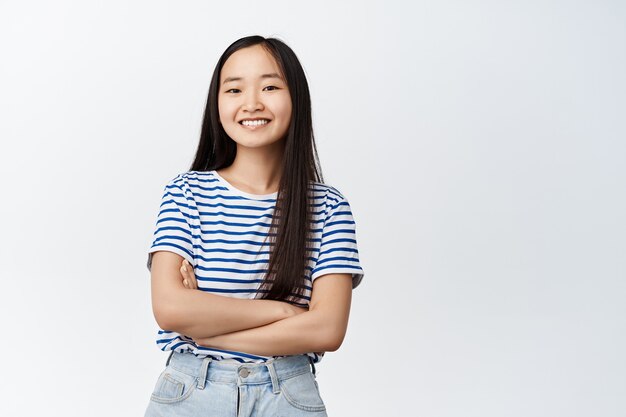 Lächelndes asiatisches Mädchen, das glücklich aussieht, die Arme auf der Brust verschränkt und im gestreiften T-Shirt auf weiß steht