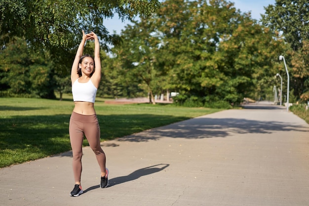 Lächelndes asiatisches Mädchen, das sich nach einem guten Training im Park ausdehnt und Musik in drahtlosen Kopfhörern joggt