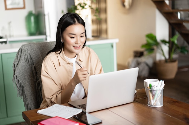 Lächelndes asiatisches Mädchen, das zu Hause auf dem Laptop lernt, das Webinar hört und Notizen macht, die Hausaufgaben aufschreiben ...