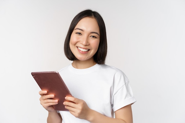 Lächelndes asiatisches Mädchen mit digitalem Tablet, das glücklich und lachend in T-Shirt auf weißem Hintergrund posiert