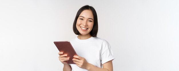 Lächelndes asiatisches Mädchen mit digitalem Tablet, das glücklich und lachend in T-Shirt auf weißem Hintergrund posiert