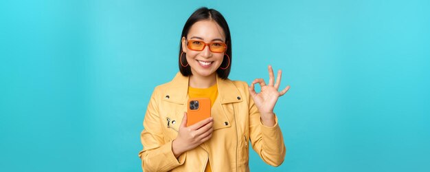 Lächelndes asiatisches Mädchen mit Smartphone, das zeigt, dass es okay ist, sich anzumelden, wenn es auf blauem Hintergrund steht