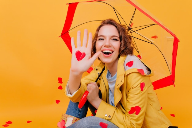 Lächelndes bezauberndes mädchen, das papierherz hält, während unter regenschirm sitzend. innenfoto der dunkelhaarigen frau, die am valentinstag aufwirft.