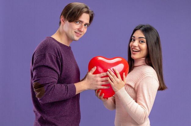 Lächelndes Blick in die Kamera junges Paar am Valentinstag mit Herzballon isoliert auf blauem Hintergrund