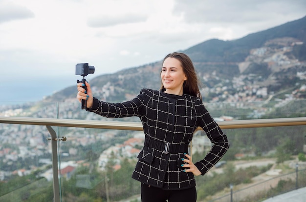 Lächelndes Blogger-Mädchen macht Selfie mit ihrer Kamera, indem sie vor dem Hintergrund der Stadtansicht die Hand auf die Taille legt