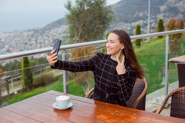 Lächelndes Bloggermädchen macht Selfie mit Smartphone, indem es zwei Gesten zeigt, indem es auf dem Hintergrund der Stadtansicht sitzt