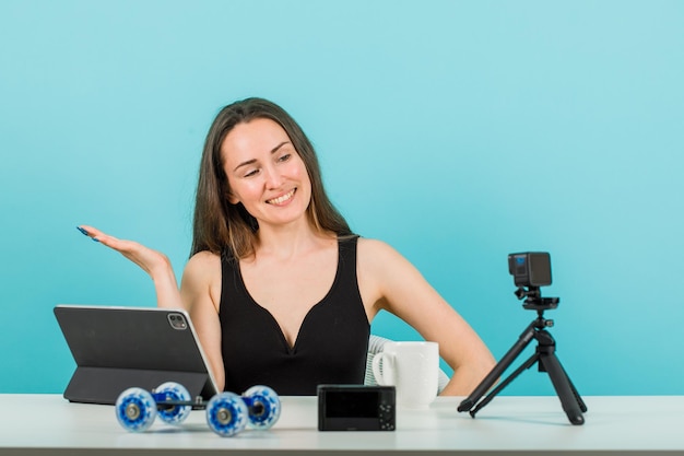 Lächelndes Bloggermädchen schaut auf ihre kleine Kamera, indem sie mit der Hand auf blauem Hintergrund nach links zeigt