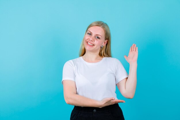 Lächelndes blondes Mädchen hebt ihre Handvoll auf blauem Hintergrund auf