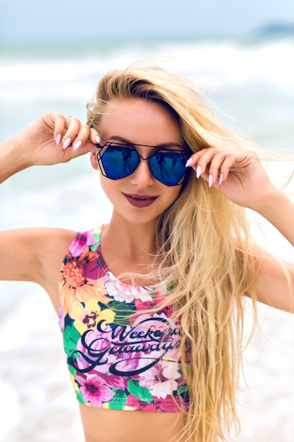 Lächelndes blondes Mädchen in der dunklen Sonnenbrille, die auf Seehintergrund aufwirft. Außenporträt der blonden Frau im Trägershirt mit Blumendruck, der im Ozeanresort im Sommer entspannt.
