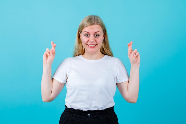 Lächelndes blondes Mädchen schaut in die Kamera, indem es die Finger auf blauem Hintergrund kreuzt