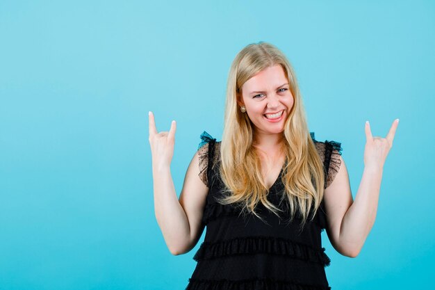 Lächelndes blondes Mädchen schaut in die Kamera, indem es Rockgesten auf blauem Hintergrund zeigt