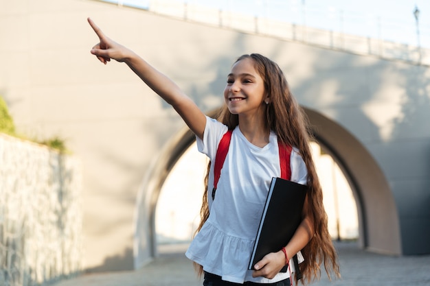 Lächelndes brünettes Schulmädchen, das draußen zeigt und wegschaut