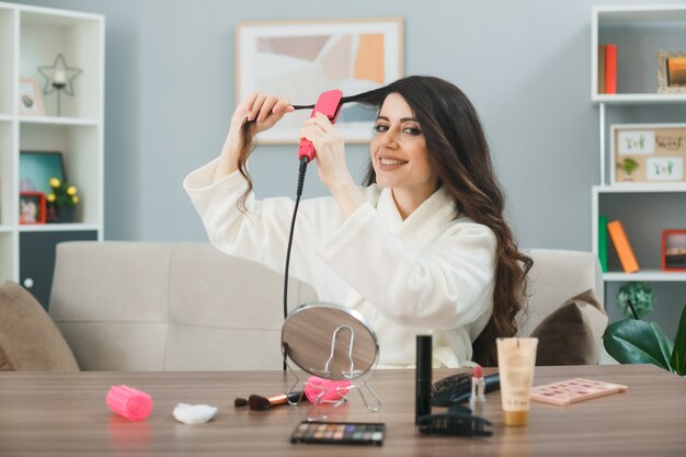 Lächelndes glätten Haar mit flachem Eisen junges Mädchen sitzt am Tisch mit Make-up-Tools im Wohnzimmer