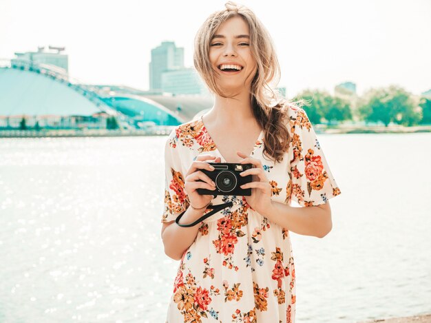 lächelndes Hipster-Mädchen im trendigen Sommer-Sommerkleid, das Retro-Kamera hält