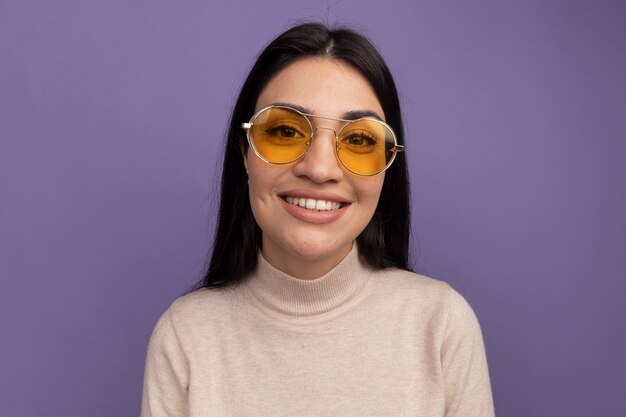 Lächelndes hübsches brünettes kaukasisches Mädchen in der Sonnenbrille betrachtet Kamera auf lila