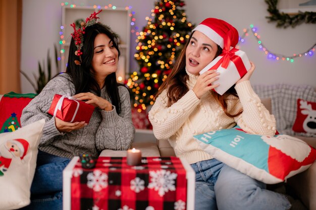 Lächelndes hübsches junges Mädchen mit Stechpalmenkranz hält Geschenkbox und sieht ihre Freundin mit Weihnachtsmütze auf einem Sessel sitzen und genießt die Weihnachtszeit zu Hause