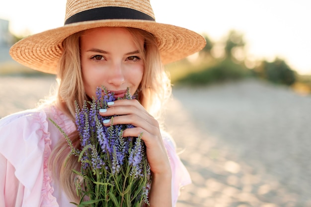 Lächelndes hübsches Mädchen im Strohhut, das auf sonnigem Strand nahe Ozean mit Blumenstrauß aufwirft. Nahaufnahme Porträt.