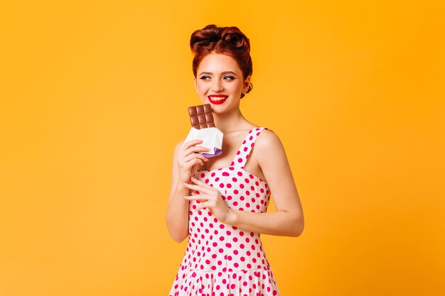 Lächelndes Ingwermädchen, das Schokolade isst. Studioaufnahme der Pinup-Frau im gepunkteten Kleid lokalisiert auf gelbem Raum.