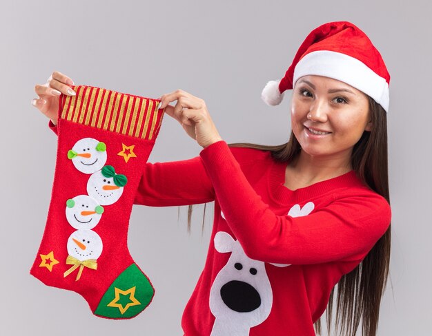 Lächelndes junges asiatisches Mädchen, das Weihnachtsmütze mit Pullover hält Weihnachtssocke lokalisiert auf weißer Wand trägt