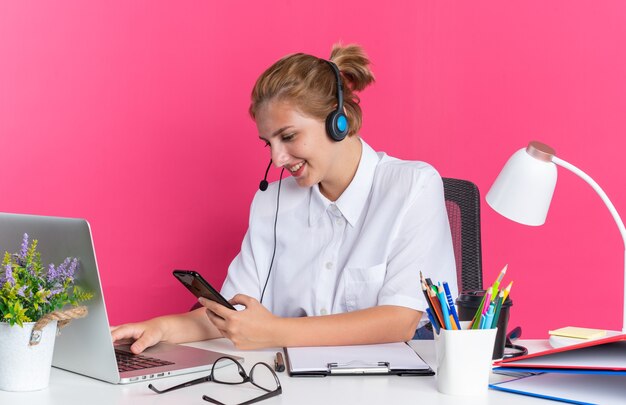 Lächelndes junges blondes Call-Center-Mädchen mit Headset am Schreibtisch sitzend mit Arbeitswerkzeugen mit Laptop und Mobiltelefon isoliert auf rosa Wand