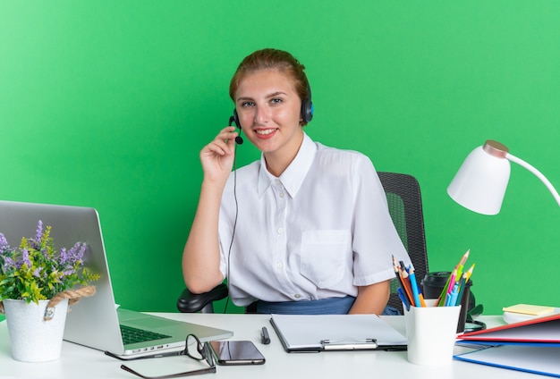 Lächelndes junges blondes Callcenter-Mädchen mit Headset am Schreibtisch sitzend mit Arbeitswerkzeugen, die das Headset-Mikrofon greifen