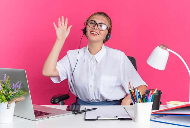 Lächelndes junges blondes Callcenter-Mädchen mit Headset und Brille, das am Schreibtisch mit Arbeitswerkzeugen sitzt und auf die Seite schaut, die Hi-Geste einzeln auf rosa Wand macht?