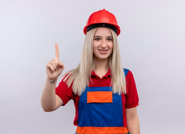 Lächelndes junges blondes Ingenieur-Baumeistermädchen in der Uniform in Zahnspangen, die Finger auf lokalisierten weißen Raum anheben