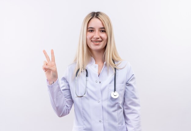 Lächelndes junges blondes Mädchen des Doktors, das Stethoskop und medizinisches Kleid in der Zahnspange trägt, die Friedensgeste auf lokalisiertem weißem Hintergrund zeigt