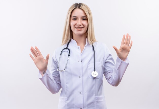 Lächelndes junges blondes Mädchen des Doktors, das Stethoskop und medizinisches Kleid in der Zahnspange trägt, die Hände auf lokalisiertem weißem Hintergrund anhebt