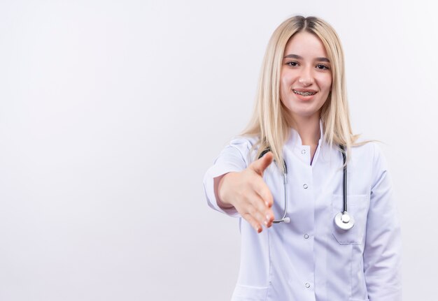 Lächelndes junges blondes Mädchen des Doktors, das Stethoskop und medizinisches Kleid in der Zahnspange trägt, die Hand zur Kamera auf lokalisiertem weißem Hintergrund heraushält