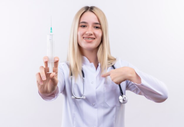Lächelndes junges blondes Mädchen des Doktors, das Stethoskop und medizinisches Kleid in Zahnspange hält, die Spritze hält, zeigt auf sich auf lokalisiertem weißem Hintergrund