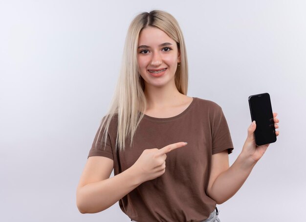 Lächelndes junges blondes Mädchen in Zahnspangen, die Handy halten und auf isolierten weißen Raum mit Kopienraum zeigen