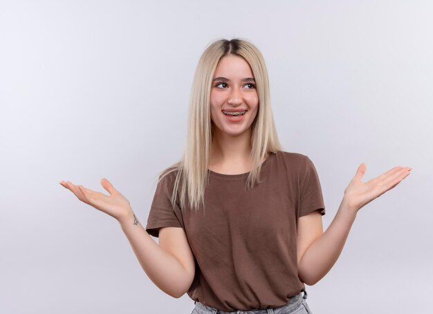 Lächelndes junges blondes Mädchen in Zahnspangen, die leere Hände auf lokalisiertem Leerraum zeigen