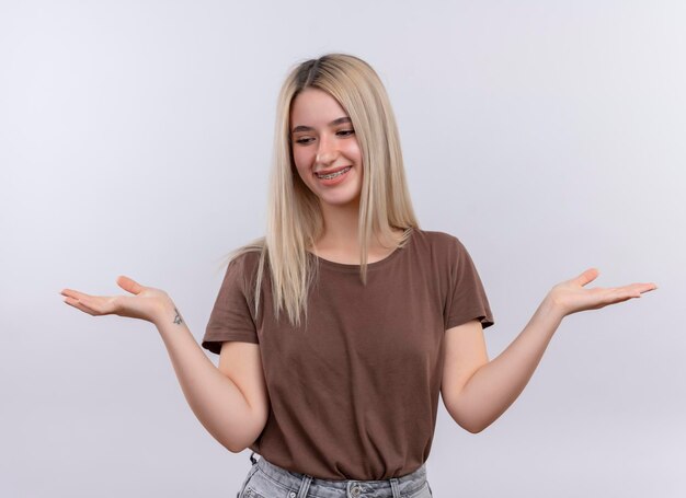Lächelndes junges blondes Mädchen in Zahnspangen, die leere Hand auf lokalisiertem weißen Raum zeigen