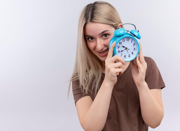 Kostenloses Foto lächelndes junges blondes mädchen in zahnspangen, die wecker auf lokalisiertem leerraum mit kopienraum halten