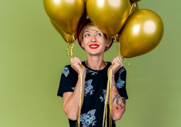 Lächelndes junges blondes Partei-Mädchen, das Ballons hält, die Seite lokalisiert auf olivgrünem Hintergrund mit Kopienraum betrachten