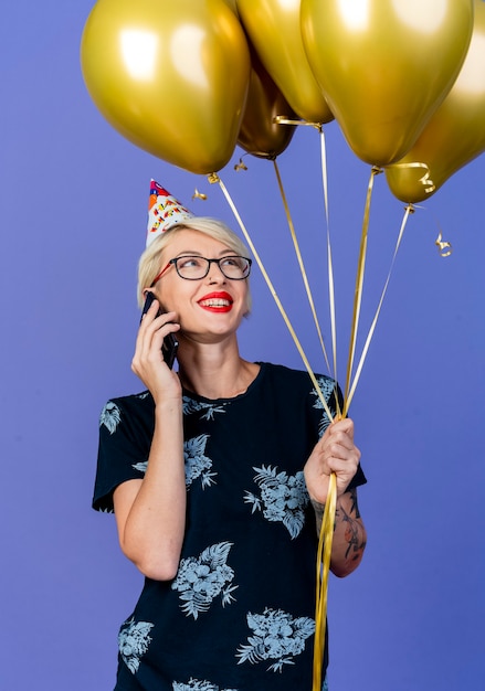 Lächelndes junges blondes Partei-Mädchen, das Brille und Geburtstagskappe hält und Ballons spricht, die am Telefon lokalisiert auf lila Hintergrund halten