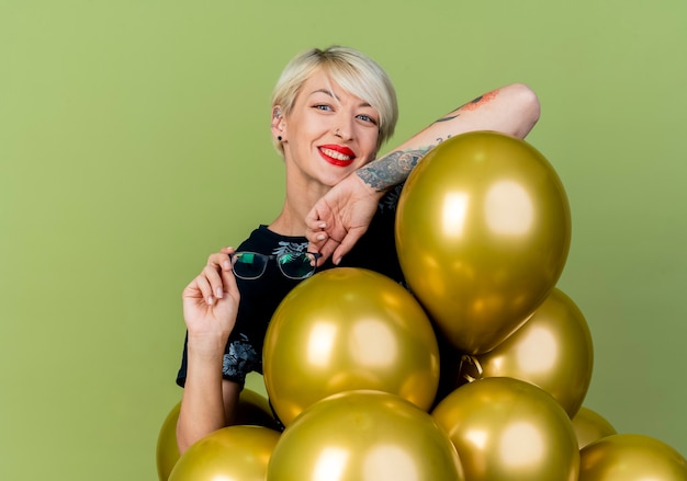 Lächelndes junges blondes Partei-Mädchen, das hinter Ballons steht, die Gläser halten Arm auf Ballon setzen betrachten Kamera lokalisiert auf olivgrünem Hintergrund mit Kopienraum