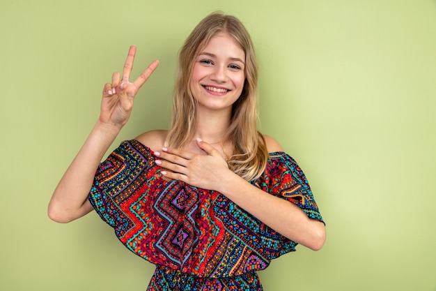 Lächelndes junges blondes slawisches Mädchen, das Hand auf ihre Brust legt und Victory-Zeichen gestikuliert