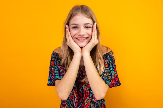 Lächelndes junges blondes slawisches Mädchen, das sich die Hände aufs Gesicht legt und nach vorne schaut