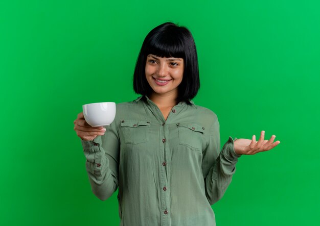 Lächelndes junges brünettes kaukasisches Mädchen hält Tasse und betrachtet Seite lokalisiert auf grünem Hintergrund mit Kopienraum