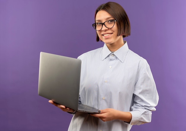 Lächelndes junges Callcenter-Mädchen, das Brillen hält Laptop lokalisiert auf lila Wand